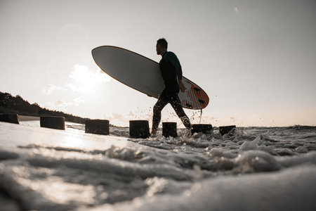 Handsome young male surfer walks along the shore past wooden posts and holds a surfboard in his...