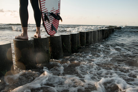 Male surfer legs in wetsuit standing on wooden stump near surfboard close up