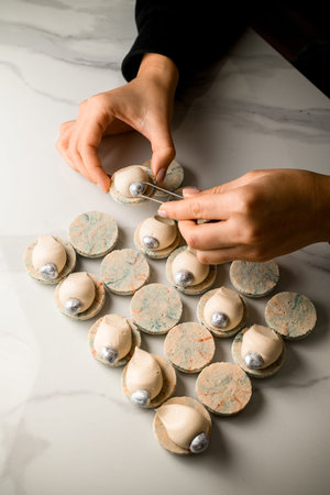 Top view of confectioners female hands decorating the last white macaron with a silver nut like a...