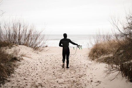 Young male surfer, dressed in a wetsuit, walks on the sand between the bushesの写真素材
