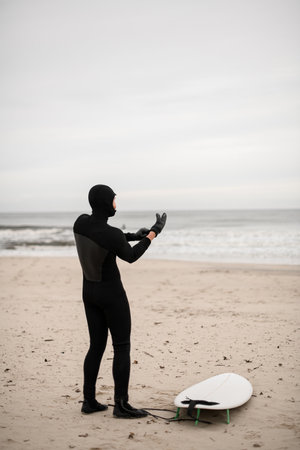 Male surfer standing on the seashore next to his surfboard and wearing surfing glovesの写真素材