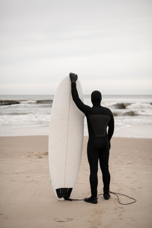 Male surfer in black wetsuit standing on sandy seashore with white surfboard standing on sandの写真素材