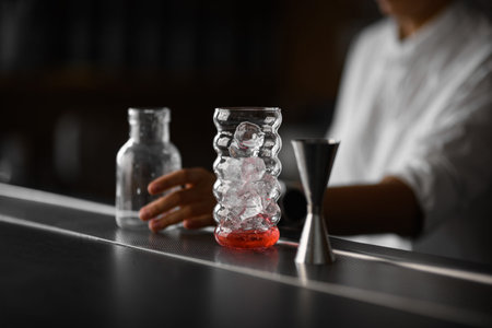 Focus on a shaped glass filled with ice cubes, next to which on the bar stand a jigger and a jar...