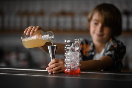 Focus on a bottle of juice that a little boy pours into a jigger to make a fruit cocktail with ice