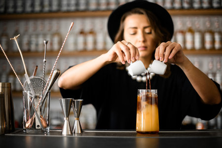 Cute female bartender pouring cinnamon drink from two white cups into tall glass with juiceの写真素材