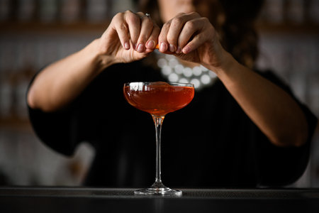 Female hands of a bartender squeezing a piece of zest, above a glass filled with a red-brown cocktailの写真素材