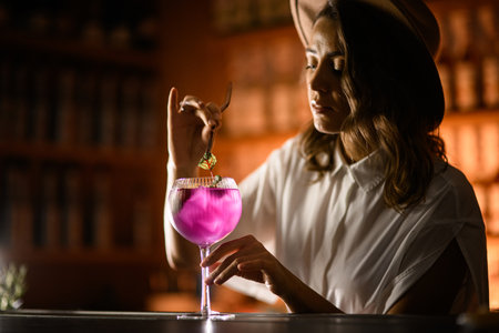 Cute female bartender in a hat prepares a purple cocktail with ice in a tall stemmed glassの写真素材