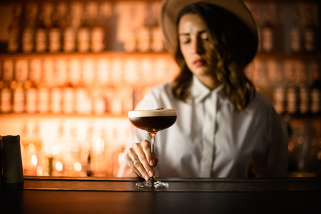 Female bartender holding cocktail glass filled with cold coffee drink with foam