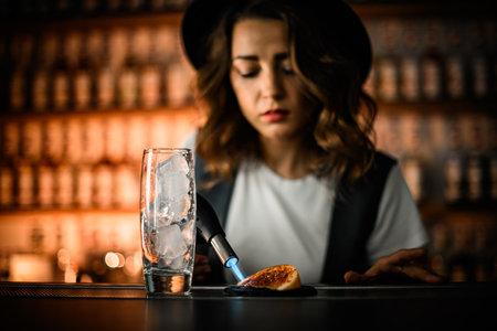 Tall glass with ice, in the background a female bartender is frying an orange sliceの写真素材