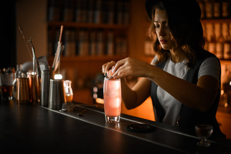 Female bartender puts a fried orange slice on a glass with a pink iced cocktailの写真素材