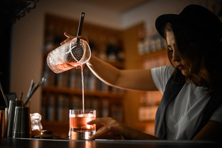 Female bartender pours a brown cocktail from a mixing glass held high above the bar into a cocktail glassの写真素材