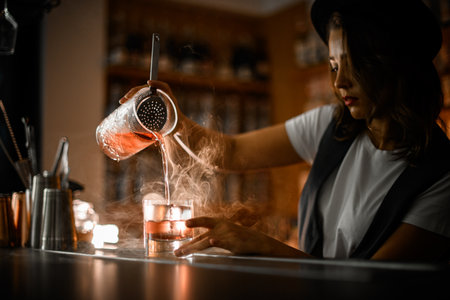 Woman bartender in a white t-shirt and vest pours a cocktail with ice from a mixing glass into another glassの写真素材