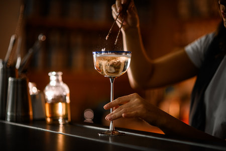 Female hands of a bartender drowned a piece of cotton candy in a glass on a tall metal legの写真素材