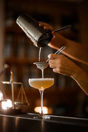 Female bartender finishes straining through two sieves a frothy champagne-colored cocktail into a stemmed glassの写真素材