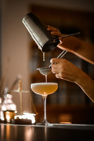 Female bartender finishes straining foamy champagne-colored cocktail prepared in a shaker through two sievesの写真素材