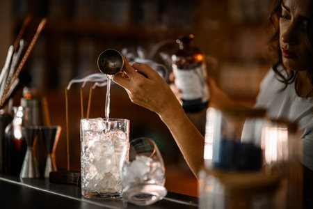 Female bartender pours a clear drink from a jigger into a mixing glass filled with iceの写真素材