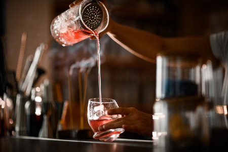 Female bartender pours a pink cocktail with ice into a tumbler glass of an unusual shapeの写真素材