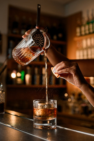 On the bar stands a glass with an ice cube, into which female hands pour a brown cocktail from a mixing glassの写真素材