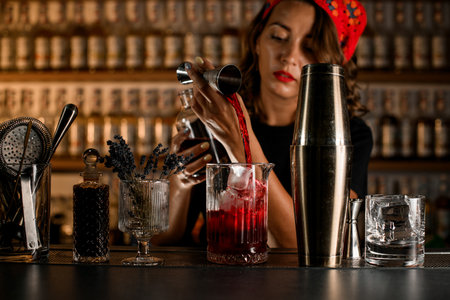 Cute female bartender pouring red wine from jigger into cocktail mixing glass with ice cubesの写真素材