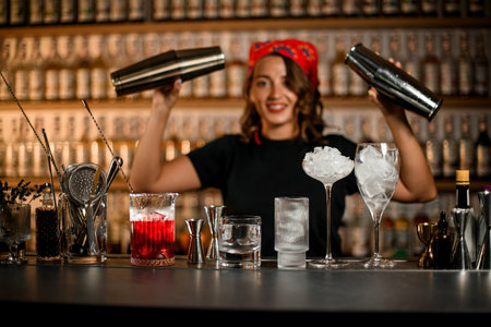 Happy smiling female bartender holding two cocktail shakers in front of various glassesの写真素材