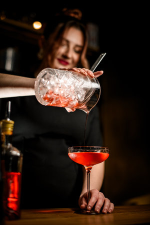 Female bartender pouring a thin stream of brown cocktail with ice from a mixing glass into a stemmed glassの写真素材