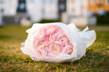 Focus on bouquet of light pink peonies in white wrapping paper lying on green grass