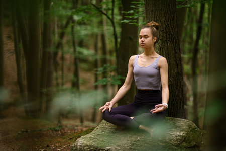 Selective focus of young woman meditating in the forest, sitting on a huge stone in the lotus positionの写真素材