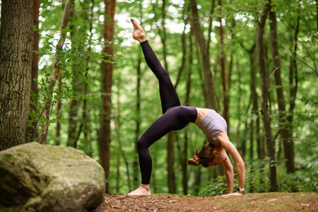 Young slender woman standing in the forest on the ground in a bridge pose with a straight leg raised...