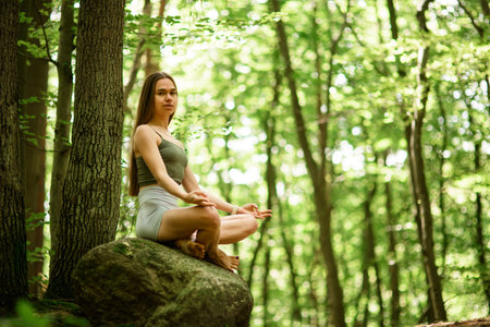 Young slim woman with long hair sitting on a big stone in a lotus position and looking at the cameraの写真素材
