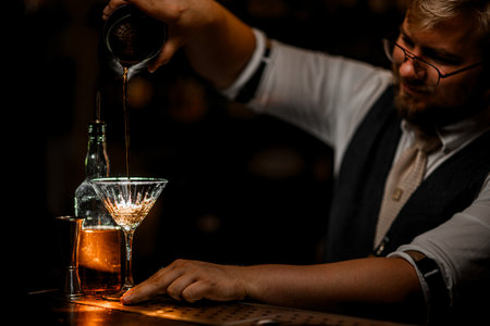 Side view of smiling male bartender pouring cocktail prepared in mixing glass into stemmed glassの写真素材