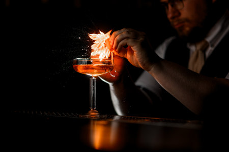 The bartender holds an orange peel over a tall stemmed glass with a cocktail, flames sparkの写真素材