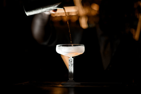 Focus on frosted stemmed glass in which a mans hand pours a cocktail from a glassの写真素材