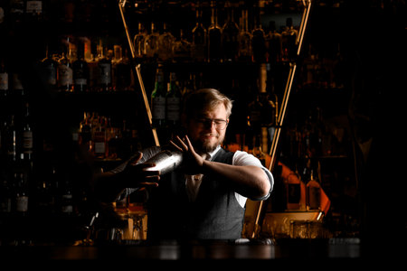 Male bartender carefully mixes a cocktail in a shaker, which he holds with two hands with his eyes closedの写真素材