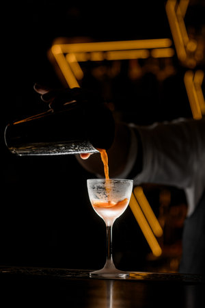Male bartenders hand pours an orange drink from a shaker into a glass on a tall stem, covered with...