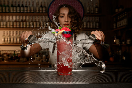 Female bartender pours water from two jugs into a glass with a pink cocktailの写真素材