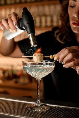 Female bartender holds a wooden skewer with a piece of banana strung with sugar sprinkled over a glass of drinkの写真素材