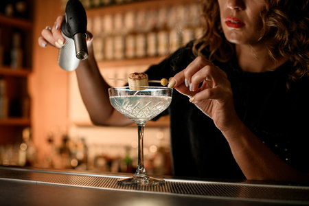 Female bartender holds a wooden skewer with a piece of banana strung on it and melted sugar and a portable gas burnerの写真素材