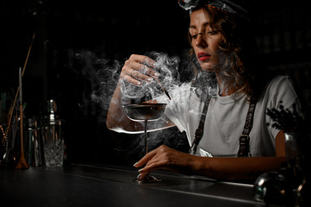 Female bartender looking through smoke at stemmed glass, blowing smokeの写真素材