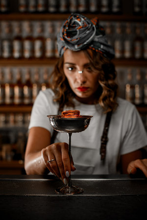 Female bartender holds the stem of a tall glass with a cocktail in front of her and looks intently...