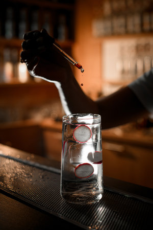 A hand in the dark drips drops with a pipette into a glass with a clear iced cocktailの写真素材