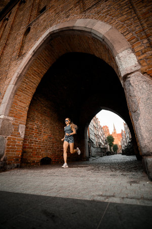 Young athletic woman in sportswear, in a fast intense pace, runs out of an archway between houses