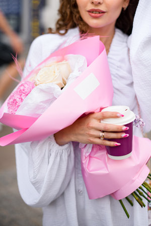 Pretty woman in a white dress is holding a cardboard cup and a bouquet of flowersの写真素材