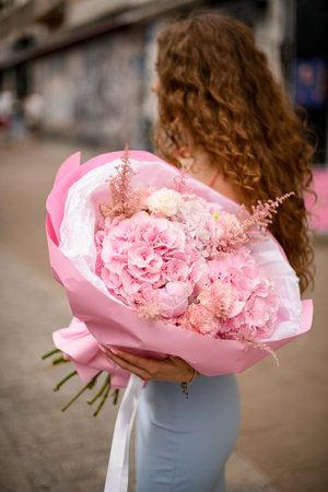 Girl with charming curly hair stands on a street with bouquet in handsの写真素材