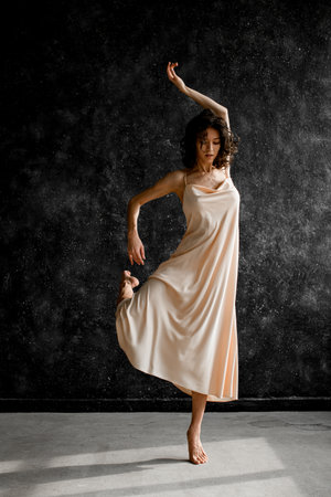 Slim woman in a long pink dress is dancing ballet, raising one arm and bringing back one legの写真素材