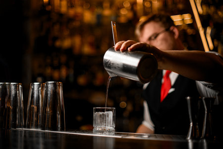 Selective focus on glass tumbler with ice cube into which bartender pours clear cocktailの写真素材