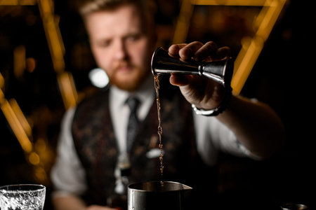 Bartenders hand pours a thin stream of liquid from a jigger into a metal glass