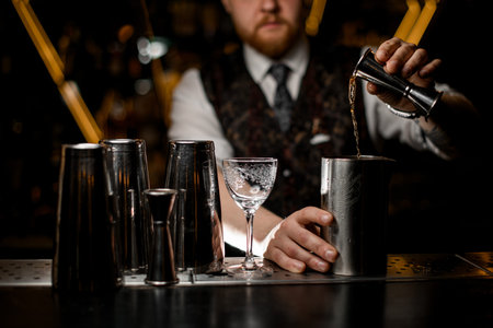 Selective focus on male bartenders hand pours liquid from cocktail jigger into metal glass