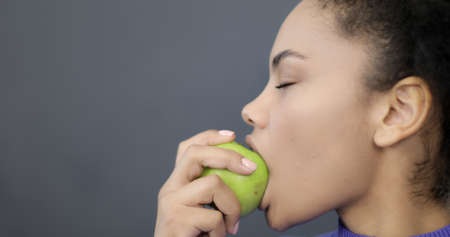 Portrait of a black woman biting a green apple. African American model eating fruit on a sofa. Healthy eating.の写真素材