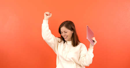 A young female student in a hoodie makes notes on a tablet on an orange background. A woman writes with a pen on a new gadget.の写真素材