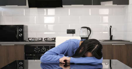 Tired young Asian woman fell asleep at the table in the kitchen. Sad multiracial girl resting at home.の写真素材